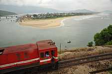 In the foreground, a red diesel locomotive is travelling up a mountain against the backdrop of Lang Co Beach and the sea. In the background are the mountains.