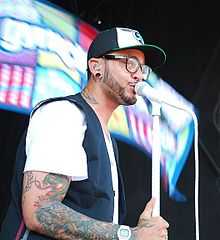 An African-American man sings into a white microphone whilst wearing a white shirt and black and white baseball cap.