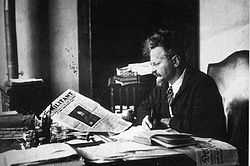 Trotsky sitting at a table opening the paper, photographed from the side