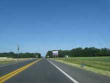 A two-lane road passing through farm fields.  A blue and white sign on the right indicates an ongoing construction project on US 113