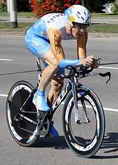A road racing cyclist in a blue, gold, and white skinsuit, sitting low in a crouch on his bicycle.