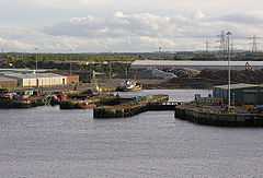 A view of a small dock as seen from a departing ferry. Industrial buildings surround, and a small boat is docked in the corner.