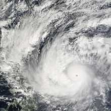 A well organized tropical cyclone, centered at bottom right, with a well-defined eye and spiraling rainbands. A landmass is visible under heavy cloud cover at left.