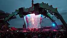 A concert stage; four large legs curve up above the stage and hold a video screen which is extended down to the band. The legs were lit up in green. The video screen has multi-coloured lights flashing on it. The audience surrounds the stage on all sides.