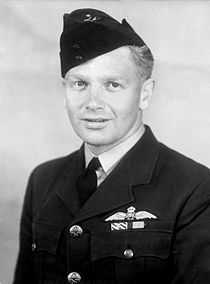 Formal head-and-shoulders portrait of man in dark military uniform with pilot's wings on breast pocket, wearing a forage cap