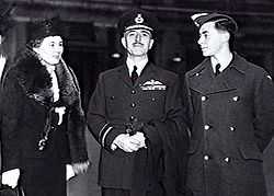 Three-quarter outdoor portrait of moustacioed man in military uniform with peaked cap and pilot's wings on left breast pocket, flanked by woman in hat and fur coat, and young man in military great coat and forage cap