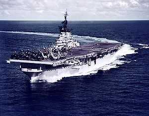 A color photo of a large aircraft carrier moving through the ocean with a deck full of aircraft