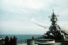 A forward view of a ship with large gun barrels pointing forward. To the left, a missile is flying away from the ship.