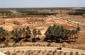 From the foot of Saddam Hussein's summer palace a Humvee is seen driving down a road towards the left. Palm trees grow near the road and the ruins of Babylon can be seen in the background.