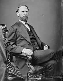 A bearded man sits in an ornate wooden chair, wile wearing a dark formal suit