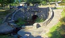 A decorative stone bridge, with curving stairs down to the water below on either side, in a park