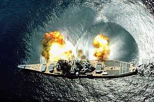 An overhead view of a large ship with a teardrop shape firing guns toward the top of the image.