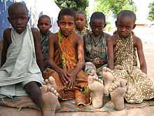 A photograph of six boys looking at the viewer and sitting on the ground with the soles of their feet facing the viewer on a bright day