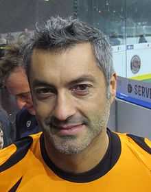 Head shot of a grinning man with brown eyes and short gray hair and trimmed gray beard. He wears an orange shirt with black-lined collar.