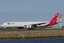 White aircraft with red tail taxiing. In the foreground is the sea. On the right is an air control tower.