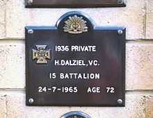 A dark plaque on a white brick wall. The plaque is decorated with a Victoria Cross emblem and reads "1936 Private H. Dalziel, VC. 15 Battalion, 24-7-1965 Age 72."