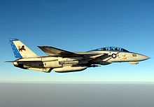 Pale gray jet aircraft flying over water towards right, perpendicular to the camera. Horizon located two-thirds down the photo. Sky made up of two shades, dark blue covers the top, blending with a lighter shade until it is almost white above horizon