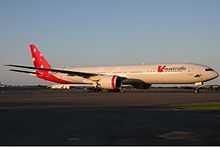 A Boeing 777-300ER in red and white livery with the Southern Cross painted on the tail. It is being parked on a sunny, cloudless day, facing right.