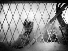 A black and white image of the film depicting the village docter's heads and arms above flour as he presses up against a chain link fence.