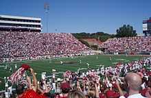 American football players line up for a kickoff on an American football field surrounded by crowded stands.