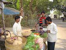A road-side vegetable-seller weighs goods while two customers check the vegetables.