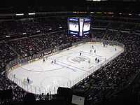 A large. white ice arena in an oval shape sits in the center of an indoor stadium which surrounds it. Fans dot the seats in white.