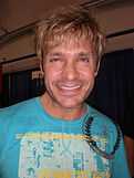 A man in a light blue t-shirt smiles towards the camera.  He has blond tousled hair, and sports a stubble.