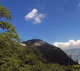 A wooded mountain with tall antennas around its summit and medium-height buildings below it. There is a tree on the left of the image, and on the right the mountain slopes down to an area next to water with some taller buildings.