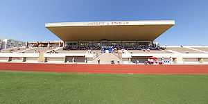 The stand of a football stadium, appearing to be made of concrete, in the daytime. The centre section of the stand is covered by a roof. On the front edge of the roof, the letters "VICTORIA STADIUM" can be seen.