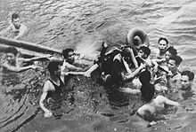 Ten men in water, surrounding one man being floated on wooden poles and tires