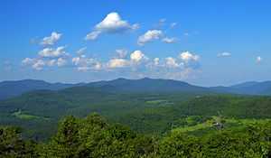 A range of mountains, mostly rounded and covered with green woods, seen past fields in the distance. Above them is a blue sky filled with little fluffy clouds.