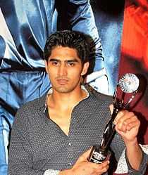 Picture of a young Indian male up to the waist. He has sharp features and short cropped black hair. He wears a black shirt and holds a trophy in his left hand