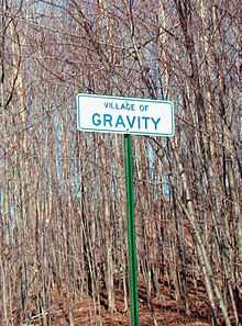 A white sign reading "VILLAGE OF GRAVITY" in green lettering, with dead trees in the background.