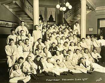 About 50 members of the 1935 freshmen class gather on the first floor of their dormitory, which has columns, a chandelier with spherical lights, and multiple staircases.