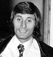A man with dark hair is wearing a white shirt with a tie and a black jacket.
