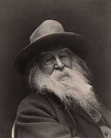 A smiling old man with a hat, dark jacket and long grey beard