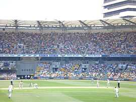 A bowler, midway through delivery. An umpire with black trousers and a light shirt watches from the left while the batsman not receiving the delivery is already walking down the pitch. In the background is a two-tier stand, full of spectators.