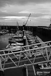 small boats lined up in harbour. Crane in the background & metal walkway in the foreground.