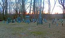 A group of grave markers, some large, some small, seen with bare trees and a dusky sky behind them