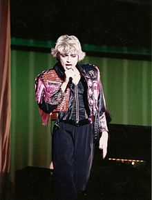 Madonna wearing a majenta jacket and black pants singing to a microphone held in her right hand