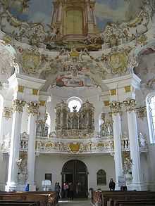 Ornate church interior, looking toward the entrance. The interior is white, the doors flanked by two pairs of columns which stretch to the richly painted ceiling. Above the entrance is the church's pipe organ.