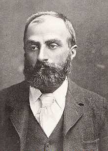  Heavily bearded man with dark receding hair, wearing a dark coloured jacket, white collar and pale tie. He is looking slightly to the left, with a solemn expression