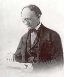 An older man with a receding hairline and reading glasses looks down at the book he is reading. He wears a dark suit with an old fashioned bow-tie