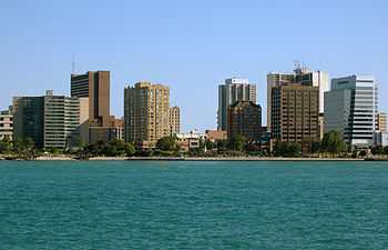 A landscape photograph of a modern North American city beside a river, taken from the other side of the water at a distance of perhaps 300 metres. A number of high-rise buildings are visible as well as a paved promenade on the waterfront.