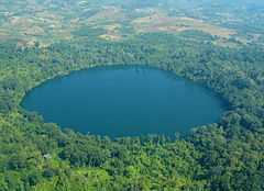 A deep blue, round lake surrounded with forest. Nearby, the forest has been replaced with fields.
