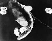 An overhead view of a large warship partway through a turn to the right. The ship's wake curves around behind her, and the surrounding sea is dotted with large areas of disturbed water and foam.