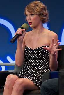 Taylor Swift speaks into a microphone, wearing a navy polka-dot dress and red heels