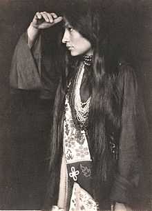 Photo of Zitkala Sa in profile, wearing Native American dress, with long dark hair hanging below waist, holding hand at forehead and looking into the distance