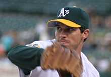 A man in a green and yellow baseball cap holds up his right hand with a baseball glove on it.