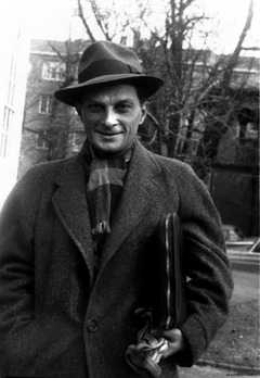 A smiling man in a hat and heavy winter coat and scarf, carrying a portfolio tucked under his arm